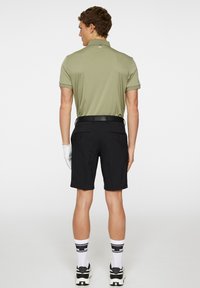 Męski strój golfowy składający się z oliwkowej koszulki polo i czarnych szortów. Szorty mają pasek, kieszenie i są zestawione z białymi skarpetami oraz trampkami.