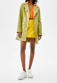 Groene glanzende jas, oranje V-hals top, gele minirok en witte sneakers. De jas heeft een reverskraag en steekzakken aan de voorkant.