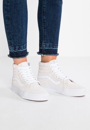 Person iført hvide højtstående sneakers med hvide snørebånd og mørkeblå opkrebede jeans mod en ensartet hvid baggrund.