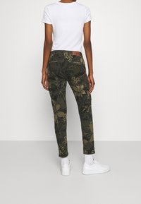 Pantalon cargo ajusté vert foncé avec motif floral, doté de deux poches latérales et d'un patch logo marron sur la ceinture. Associé à des chaussures blanches.