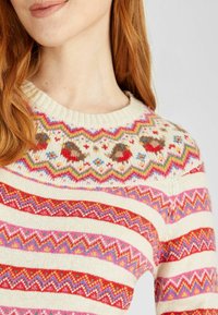 Pull tricoté avec une base crème ornée de motifs zigzag multicolores et floraux, col rond et tissu texturé.