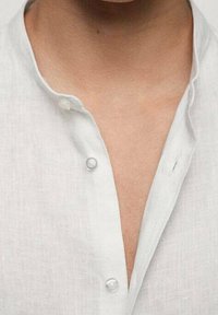 Chemise en lin blanc avec un col mandarin, dotée de deux petits boutons et d'une coupe décontractée, mettant l'accent sur des lignes épurées et une texture légère.
