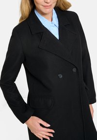 Cappotto nero doppiopetto con colletto a scollo a V, tasche frontali e due bottoni neri; indossato sopra una camicia azzurra con colletto.
