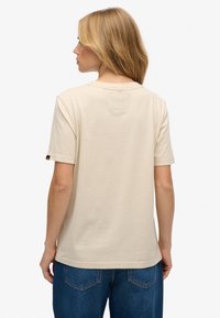 Beżowa bawełniana koszulka z krótkim rękawem, okrągłym dekoltem i małą naszywką z tyłu. Do zestawu dołączone niebieskie dżinsy.