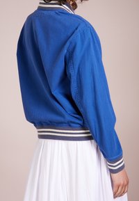 Osoba v modré bomber jacket s bílými a šedými pruhovanými manžetami a pasem, zkombinovaná s bílou plissovanou sukní.