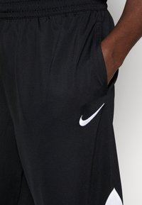 Mustat urheilushortsit, joissa on teksturoitu kangas, joustava vyötärö, sivutaskut ja valkoinen Nike-logo vasemmassa jalassa.