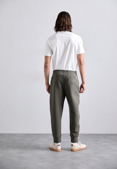 Mann med skulderlangt hår står vendt mot en vegg, iført en hvit t-skjorte, olivengrønne joggebukser, hvite sokker og beige sneakers.