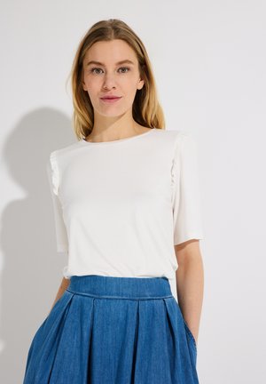 Frau mit langem blondem Haar, die ein weißes kurzärmeliges Oberteil und einen hoch taillierten blauen Faltenrock trägt, steht vor einem schlichten hellen Hintergrund.