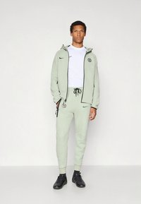 Light green Nike tracksuit with a zip-up hoodie and elastic waistband pants. Features a logo on the chest and side, paired with black shoes.