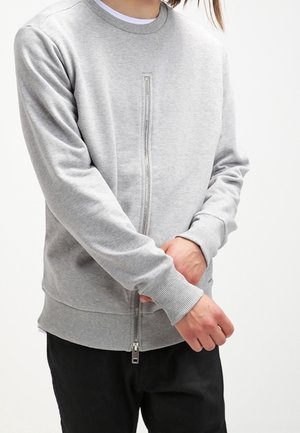 Persoon draagt een lichtgrijze zip-up sweatshirt met geribbelde manchetten en zoom, past mouw aan, gecombineerd met zwarte broek.