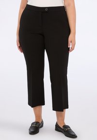 Kvinna i svarta avkortade raka byxor och svarta läder loafers med guldkedjedetalj, stående mot en vit bakgrund.