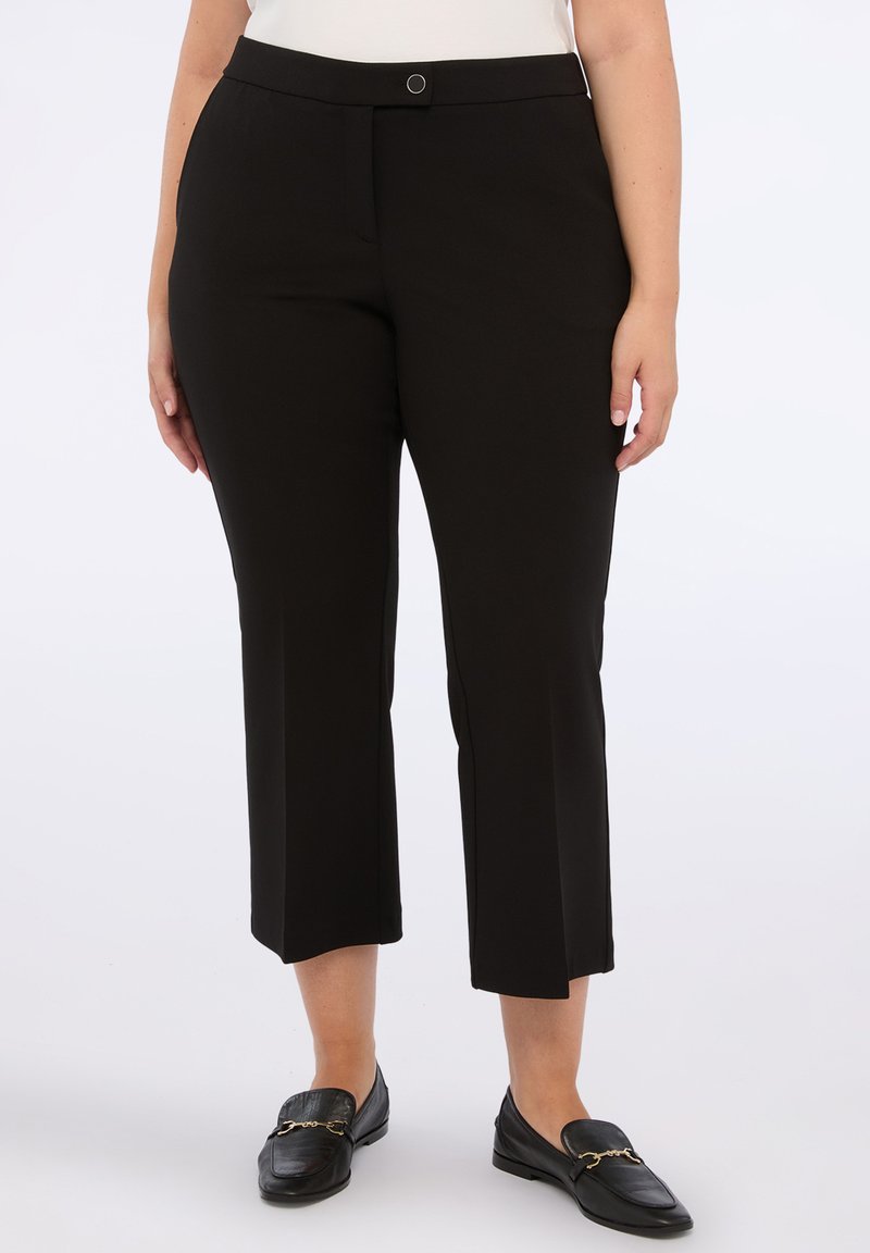 Kvinna i svarta avkortade raka byxor och svarta läder loafers med guldkedjedetalj, stående mot en vit bakgrund.