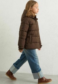 Brūns pūkains mētelis ar kapuci, ar quilted dizainu un priekšējām kabatām. Saskaņots ar zilo džinsu biksēm un brūnajām potīšu zābakiem.