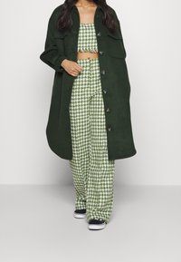 Green wool blend oversized coat over matching houndstooth crop top and wide-leg pants. Textured fabric with buttons and fringe detailing.
