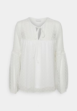 Blusa blanca hecha de tela ligera con textura de lunares. Presenta un escote en forma de ojal, mangas sueltas y detalles de encaje.