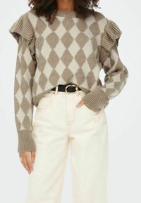 Pull à motif losanges marron et crème avec manches à volants, poignets côtelés et encolure ronde, associé à un pantalon clair taille haute et une ceinture.