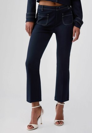 Donna che indossa pantaloni blu navy con cuciture bianche e tasche frontali, abbinati a sandali bianchi con tacco alto e cinturino alla caviglia, su sfondo semplice.