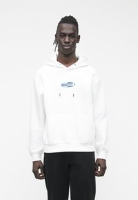 Jovem com cabelo trançado, vestido com um hoodie branco com o logotipo "Guess Jeans" e calças pretas, em frente a um fundo branco simples.
