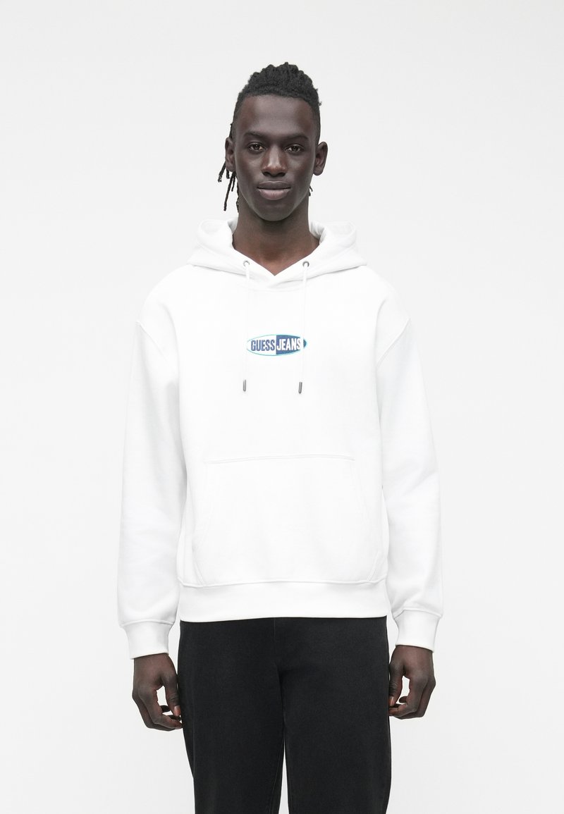 Jovem com cabelo trançado, vestido com um hoodie branco com o logotipo "Guess Jeans" e calças pretas, em frente a um fundo branco simples.
