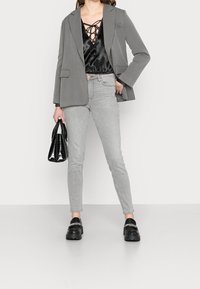 Blazer grigio sopra una blusa nera in satin con dettaglio lace-up, abbinato a jeans skinny grigi chiari e mocassini neri chunky, mentre tiene una borsa nera.