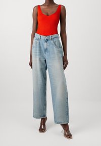 Body en maille côtelée rouge avec un décolleté en V profond, associé à un jean à taille haute et jambes larges de couleur bleu clair. Des talons blocs transparents complètent le look.