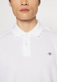 Hvid polo t-shirt med en blød tekstur, tre knaplukninger og broderet logo på venstre bryst. Klassisk krave og korte ærmer.