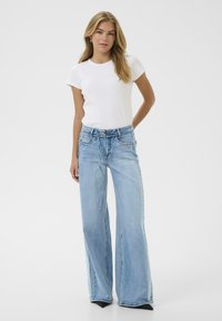 Woman standing with hand behind back wearing white short-sleeve top and light blue wide-leg jeans with black pointed shoes.