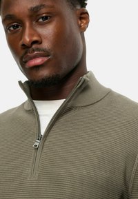 Pullover mit Viertelreißverschluss aus strukturiertem olivgrünem Stoff, mit hohem Kragen und einem silbernen Reißverschluss, über einem weißen Hemd getragen.