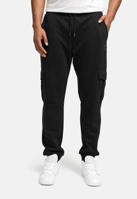 Joggers cargo negros con cintura elástica, bolsillos laterales y un corte relajado. Hechos de una suave mezcla de algodón. Combinados con zapatillas blancas.