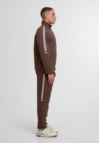 Brūna sporta jaka ar baltām svītrām, sānu kabatām un logotipu uz piedurknes, kopā ar atbilstošām biksēm un krēmkrāsas sporta apaviem.