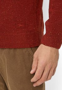 Roter Strickpullover mit strukturiertem Finish, ausgestattet mit einem gerippten Bündchen und einem kleinen Logo-Patch. Kombiniert mit braunen Cordhosen.