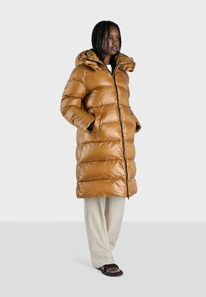 Bruine gewatteerde pufferjas met capuchon, met een ritssluiting en twee zijzakken. Draagt over een witte top en beige broek.