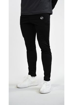 Black athletic pants made from a flexible material, featuring a snug fit, side pockets, and a silver logo. Paired with white athletic shoes.