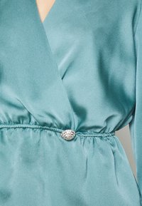 Robe en satin bleu sarcelle avec un décolleté en V profond, une taille froncée avec élastique, et un détail décoratif en broche argentée à la taille.