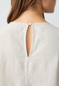 Hellbeige Leinenbluse mit einem Schlitz hinten, der mit einem kleinen, stoffbezogenen Knopf befestigt ist, und einer weichen, strukturierten Oberfläche.