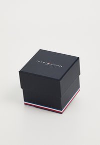 Caja cuadrada azul marino con tapa, que presenta el texto "RELOJES TOMMY HILFIGER" en blanco. La base tiene una franja tricolor en rojo, blanco y azul.