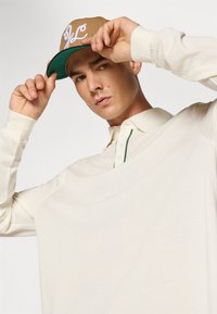 Beige langermet poloskjorte med krage og grønn detalj, brukt under en brun caps med hvit kursiv skrift.