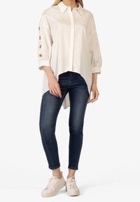 Camicia bianca con maniche a sbuffo e bottoni dorati, abbinata a jeans skinny blu scuro e sneakers beige.