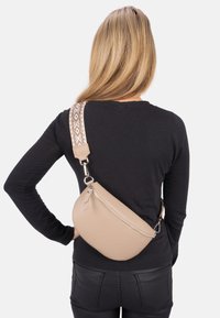 Beige Umhängetasche mit strukturierter Oberfläche, diagonalem Reißverschluss und einem gemusterten verstellbaren Trageriemen. Abgerundete Form für ergonomischen Einsatz.