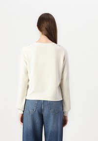 Kremkrāsas krekls ar garām piedurknēm, kuram ir noapaļota kakla izgriezums un teksturēti rotājumi uz pleciem. Apvienots ar platām zilām džinsa biksēm.