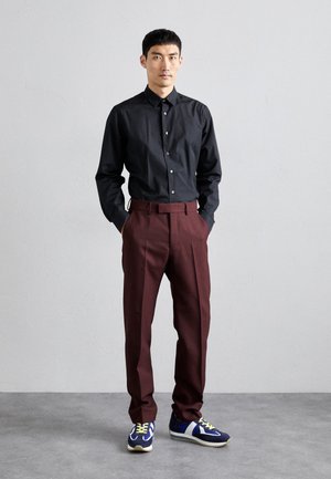 Chemise noire à manches longues et boutonnée associée à un pantalon bordeaux taille haute et des baskets bleues avec des accents verts. Le mannequin se tient avec les mains dans les poches.