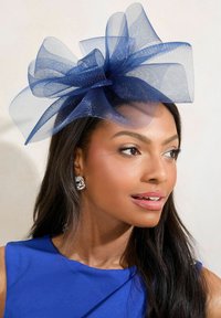 Fascinator tamnoplave boje sastavljen od slojeva tila, s velikim, prozračnim mašnom na vrhu. Model nosi srebrne naušnice s okruglim zupcima i plavu haljinu.