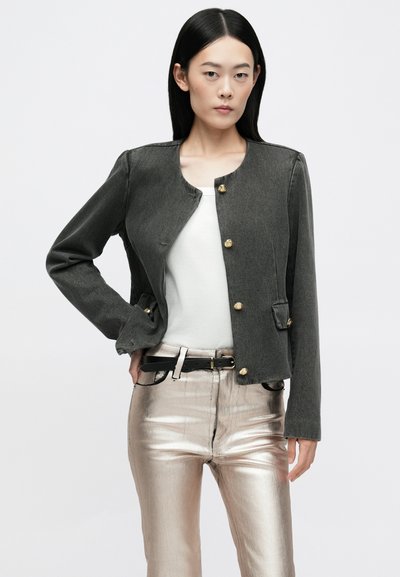 Veste en denim gris foncé avec un col rond, des boutons dorés et des poches latérales, associée à un pantalon métallique argenté, comprenant une ceinture noire.