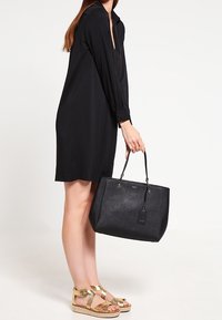 Svart tote bag med en strukturerad form och texturerad yta, med dubbla handtag och en hängande etikett, kombinerad med en svart klänning och guldsandaler.