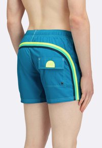 Pantaloni da bagno turchesi con striscia gialla a contrasto, vita elastica e tasca laterale con dettaglio del logo circolare.