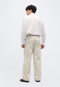 Άνδρας που στέκεται με την πλάτη προς τα εμπρός, φοράει λευκό πουκάμισο με λεπτές ρίγες, μέσα σε μπεζ παντελόνι με φαρδιά πόδια και μαύρα παπούτσια, σε απλό φόντο.