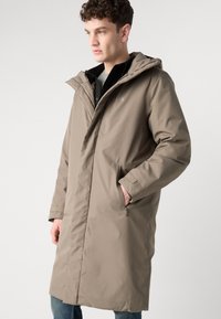Beige lang frakk med hette, med glidelås og sidelommer, laget av et glatt, vanntett materiale. CK-logo synlig på brystet.