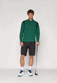 Zielona koszulka polo z długim rękawem i czarnymi akcentami, szare szorty oraz białe buty. Postawa stojąca z kijem golfowym, neutralne tło.