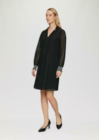 Czarna sukienka z przezroczystymi długimi rękawami, plisowanym przodem i wzorzystymi mankietami. Noszona z czarnymi szpilkami. Prosty, elegancki fason.