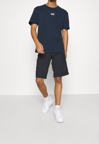 Mörkblå t-shirt med logga på bröstet, tillsammans med mörka shorts. Modellen bär vita sneakers. Enkel snitt, avslappnad tyg, minimalistisk design.
